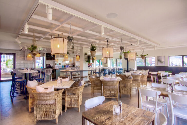 An expansive internal view showing loose timber tables, wicker armchairs, and a decorative ceiling raft.