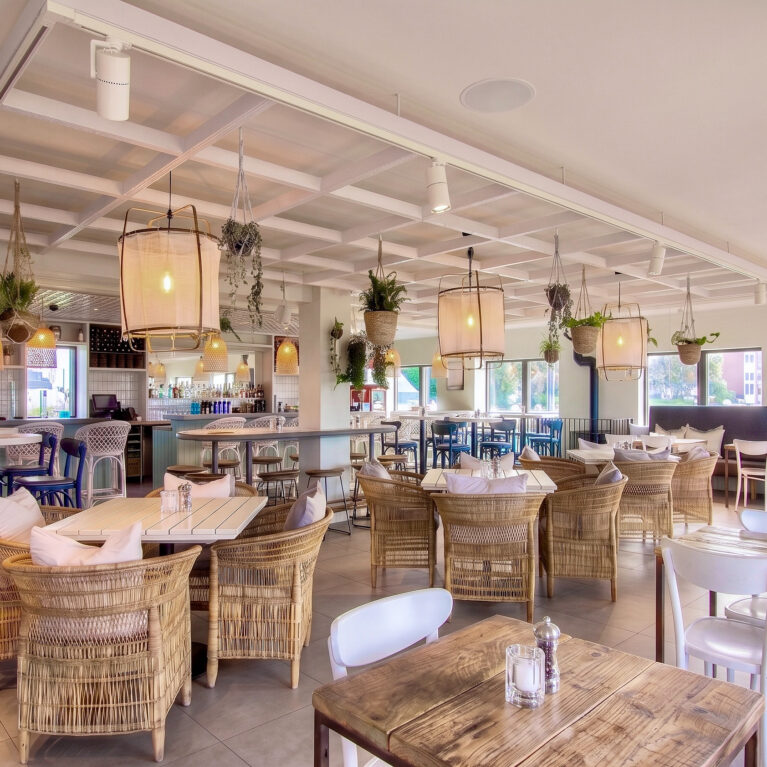 An expansive internal view showing loose timber tables, wicker armchairs, and a decorative ceiling raft.