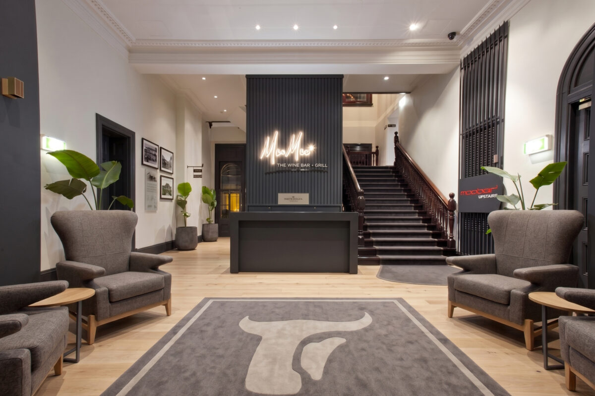 Hospitality reception desk, charcoal wall paneling, custom neon signage, heritage timber staircase, wingback lounge chairs, inlaid logo carpet, recessed ceiling downlights, commercial lobby layout.