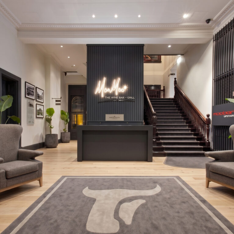 Hospitality reception desk, charcoal wall paneling, custom neon signage, heritage timber staircase, wingback lounge chairs, inlaid logo carpet, recessed ceiling downlights, commercial lobby layout.