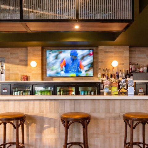 A warm, tactile composition of Safi clay tiles, engineered stone, and dark oak stools, anchored by a softly illuminated bar display.