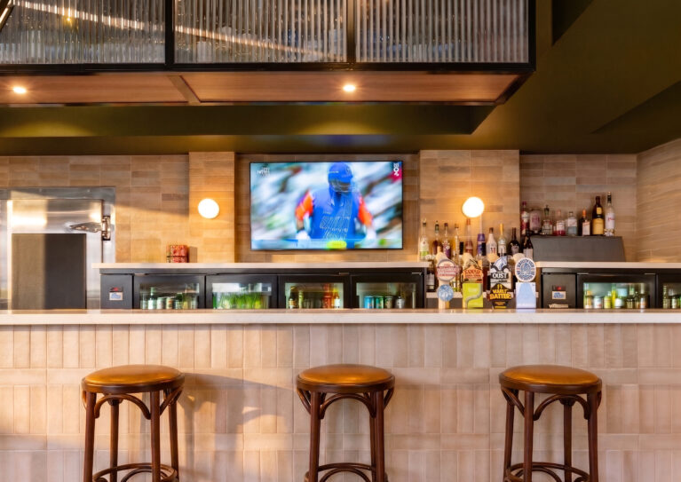 A warm, tactile composition of Safi clay tiles, engineered stone, and dark oak stools, anchored by a softly illuminated bar display.