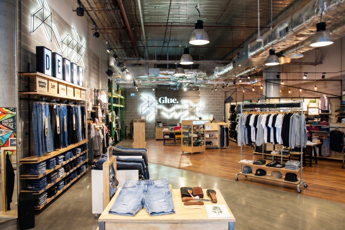An open-plan urban clothing store layout featuring exposed ceiling ductwork, a denim display wall, mixed concrete and timber flooring, and industrial pendant light fixtures