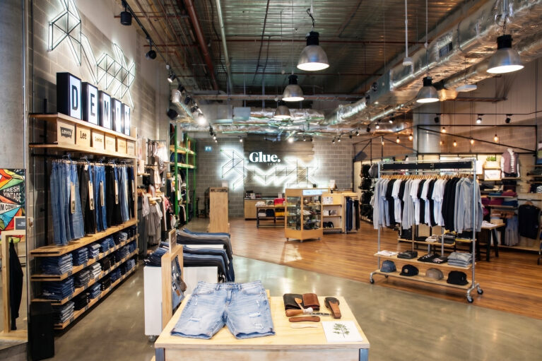An open-plan urban clothing store layout featuring exposed ceiling ductwork, a denim display wall, mixed concrete and timber flooring, and industrial pendant light fixtures