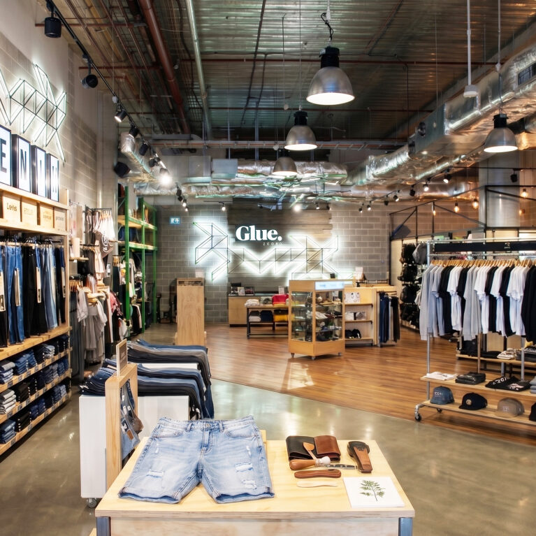 An open-plan urban clothing store layout featuring exposed ceiling ductwork, a denim display wall, mixed concrete and timber flooring, and industrial pendant light fixtures