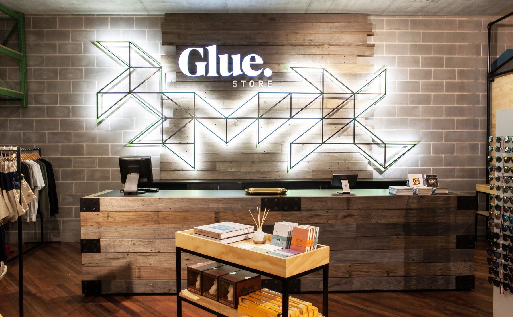 A high-end apparel retail fit-out showcasing a reclaimed timber cash wrap desk, exposed concrete block wall, geometric neon brand signage, and hardwood flooring.