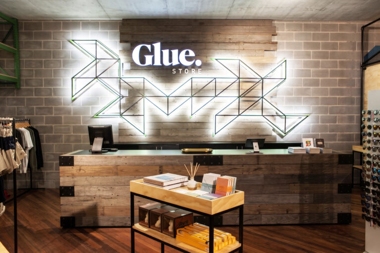 A high-end apparel retail fit-out showcasing a reclaimed timber cash wrap desk, exposed concrete block wall, geometric neon brand signage, and hardwood flooring.