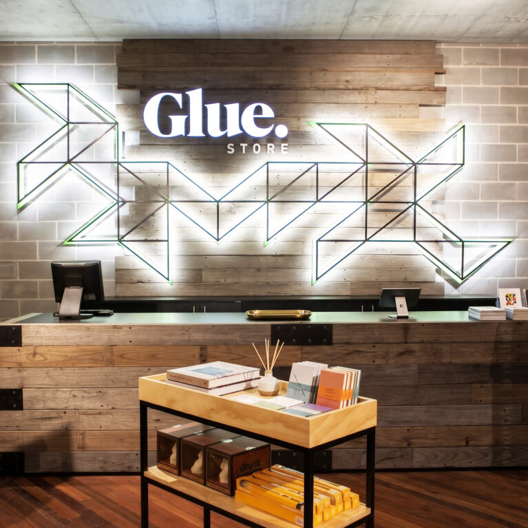 A high-end apparel retail fit-out showcasing a reclaimed timber cash wrap desk, exposed concrete block wall, geometric neon brand signage, and hardwood flooring.