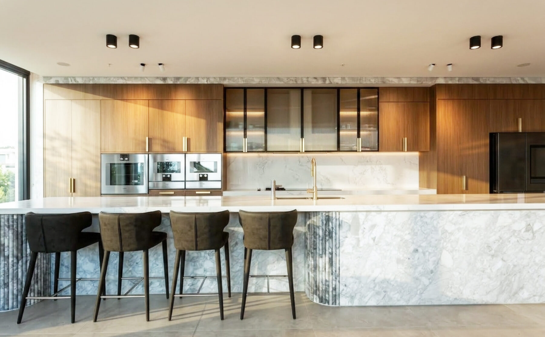 The kitchen exudes luxury and functionality, featuring a Calacatta marble island and light oak cabinetry. Black accents and integrated appliances enhance the modern aesthetic, while soft lighting highlights the refined textures of the materials.