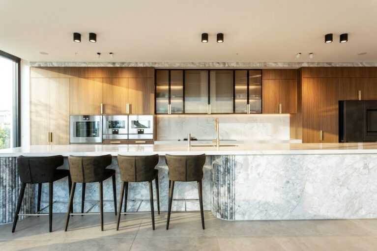 The kitchen exudes luxury and functionality, featuring a Calacatta marble island and light oak cabinetry. Black accents and integrated appliances enhance the modern aesthetic, while soft lighting highlights the refined textures of the materials.