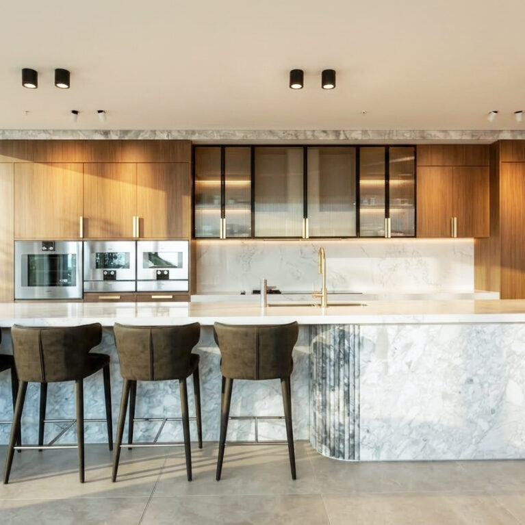 The kitchen exudes luxury and functionality, featuring a Calacatta marble island and light oak cabinetry. Black accents and integrated appliances enhance the modern aesthetic, while soft lighting highlights the refined textures of the materials.