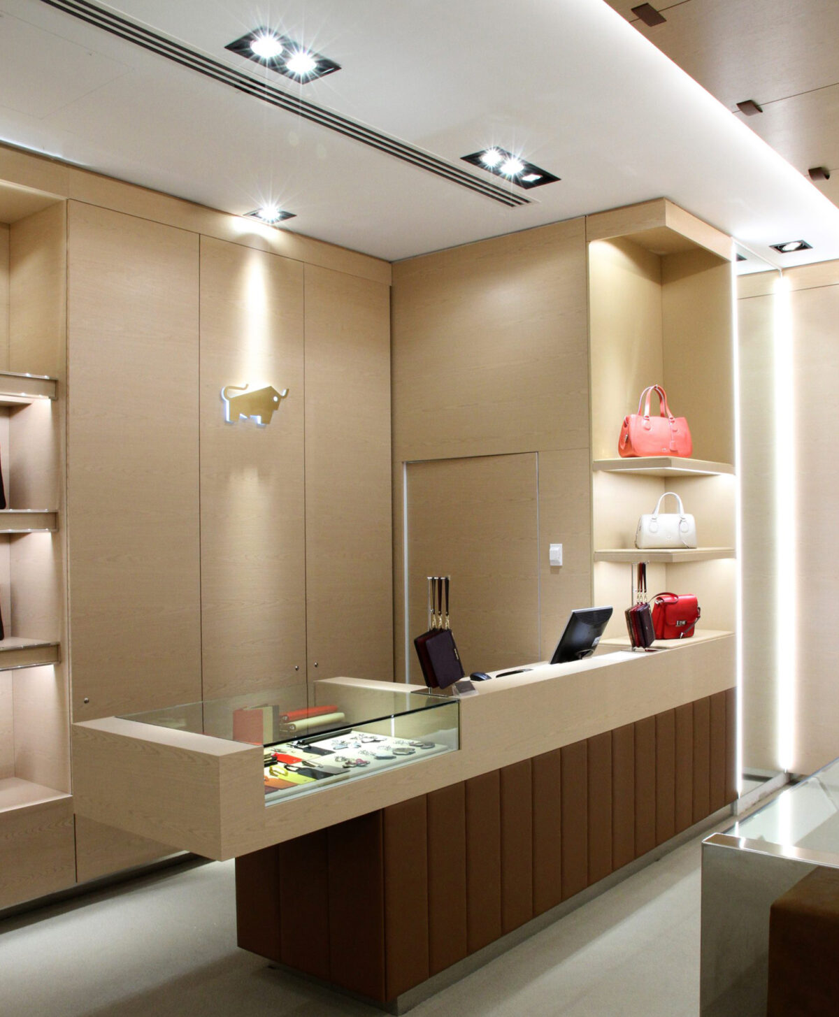 Point of sale desk, integrated glass showcase, vertical timber panels, hidden storage doors, metallic bull logo, recessed ceiling spotlights, luxury retail joinery, linear ventilation slot.
