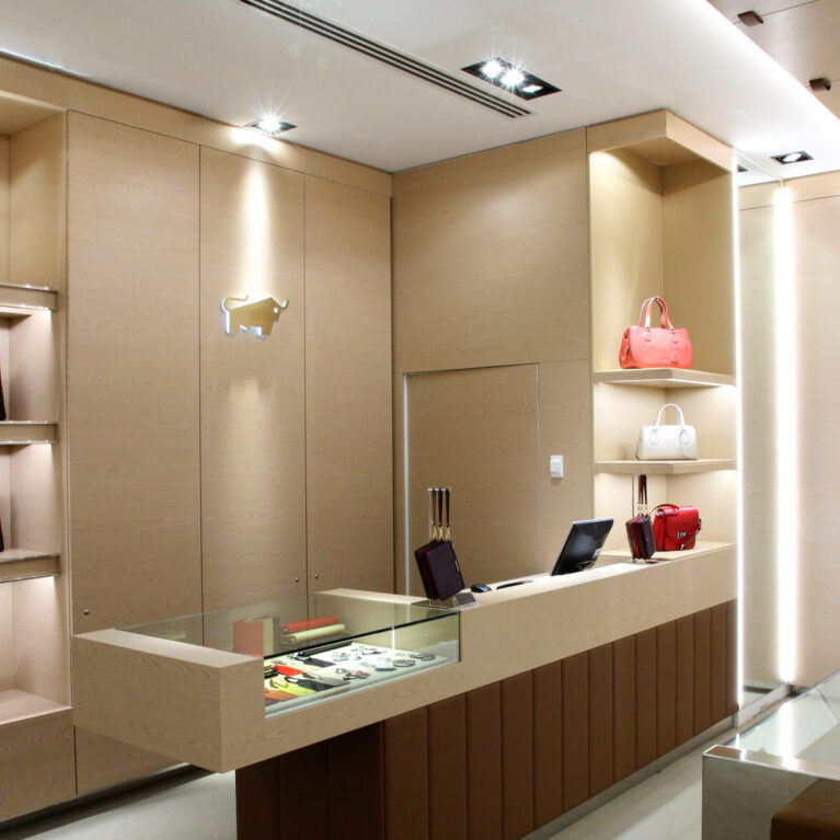 Point of sale desk, integrated glass showcase, vertical timber panels, hidden storage doors, metallic bull logo, recessed ceiling spotlights, luxury retail joinery, linear ventilation slot.