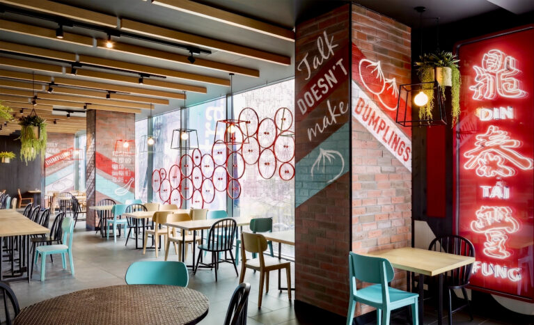Restaurant dining area, vertical neon light box, brick columns with hand-painted graphics, timber ceiling beams, wire cage pendant lamps, mixed seating layout, window decals.