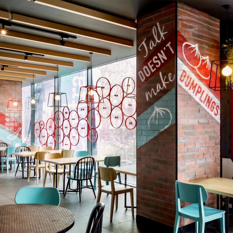 Restaurant dining area, vertical neon light box, brick columns with hand-painted graphics, timber ceiling beams, wire cage pendant lamps, mixed seating layout, window decals.