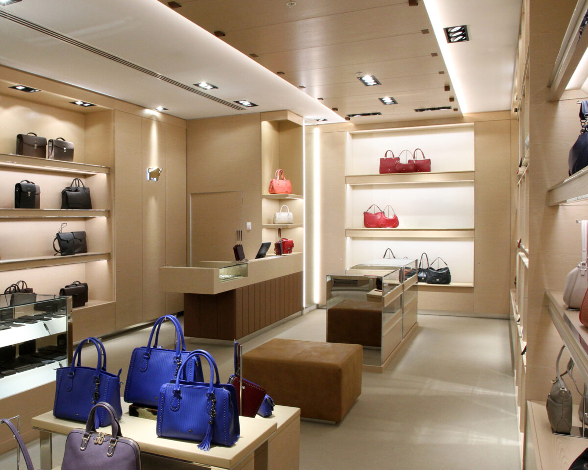 Point of sale counter, timber cladding panels, dropped ceiling bulkhead, recessed lighting design, alcove retail shelving, glass display units, leather ottoman bench, open luxury boutique layout.