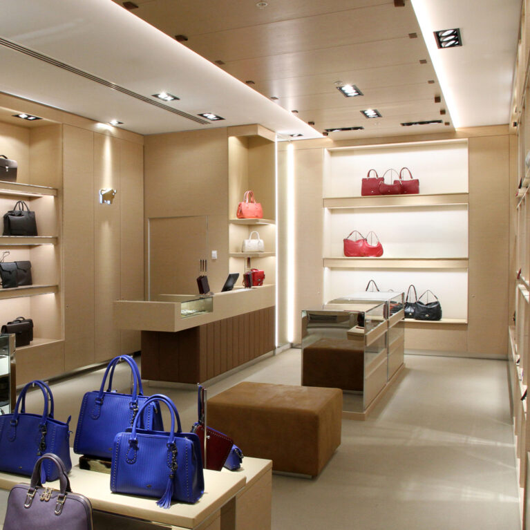 Point of sale counter, timber cladding panels, dropped ceiling bulkhead, recessed lighting design, alcove retail shelving, glass display units, leather ottoman bench, open luxury boutique layout.
