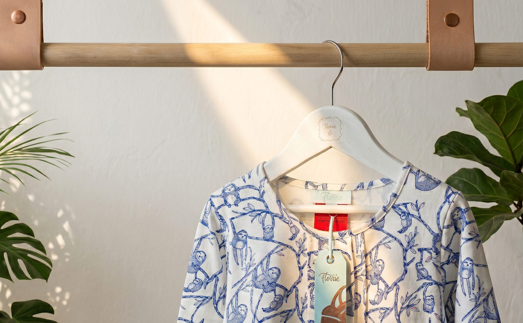 Minimalist garment display, timber dowel hanging rail, tan leather straps, white wooden clothes hanger, textured plaster wall, boutique apparel presentation, natural sunlight filtering, indoor plant framing.