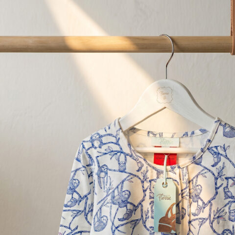Minimalist garment display, timber dowel hanging rail, tan leather straps, white wooden clothes hanger, textured plaster wall, boutique apparel presentation, natural sunlight filtering, indoor plant framing.