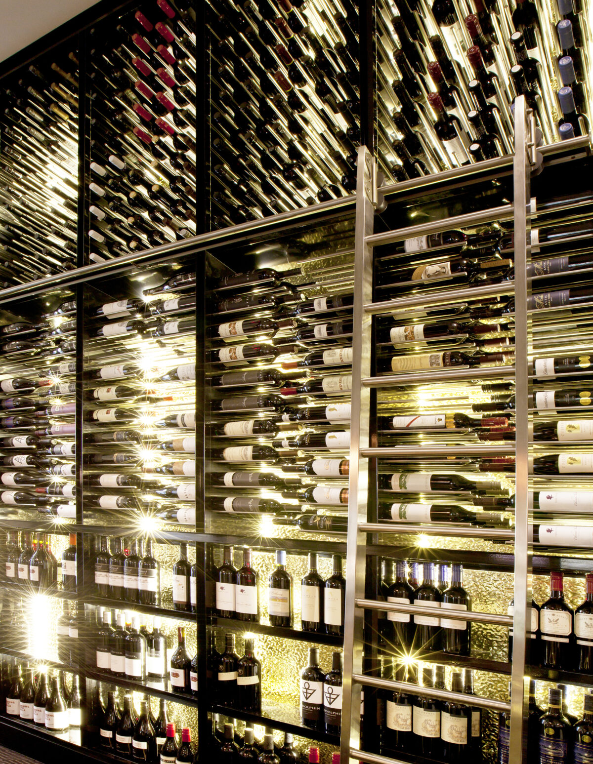 Bespoke wine display, floor-to-ceiling cellar shelves, integrated LED shelf lighting, rolling library ladder, stainless steel tracking, dark mirror backing, commercial hospitality joinery, luxury restaurant cellar.