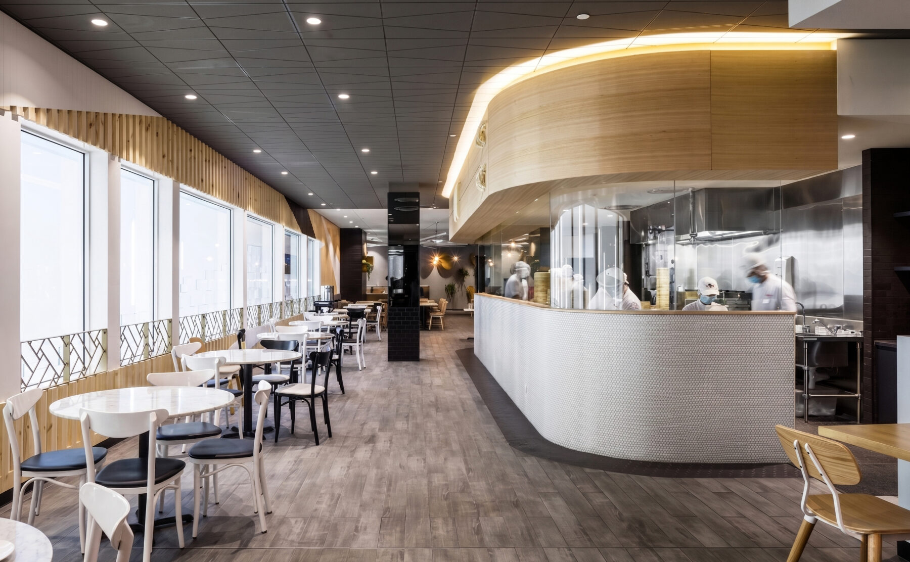 A commercial restaurant design highlighting an open show kitchen with glass panels, curved timber bulkheads, linear window seating tables, and textured grey floor tiling.