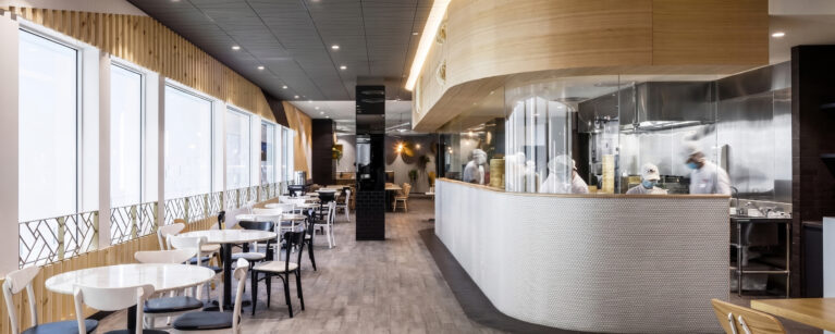 A commercial restaurant design highlighting an open show kitchen with glass panels, curved timber bulkheads, linear window seating tables, and textured grey floor tiling.
