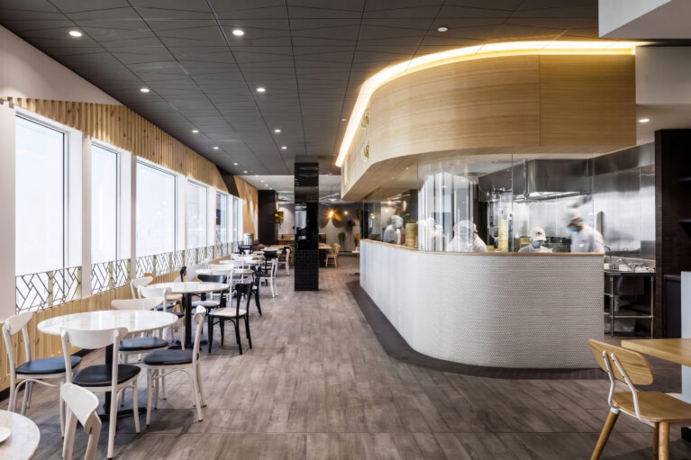 A commercial restaurant design highlighting an open show kitchen with glass panels, curved timber bulkheads, linear window seating tables, and textured grey floor tiling.