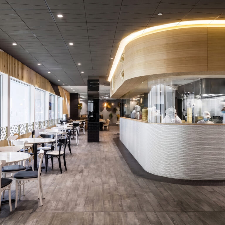 A commercial restaurant design highlighting an open show kitchen with glass panels, curved timber bulkheads, linear window seating tables, and textured grey floor tiling.