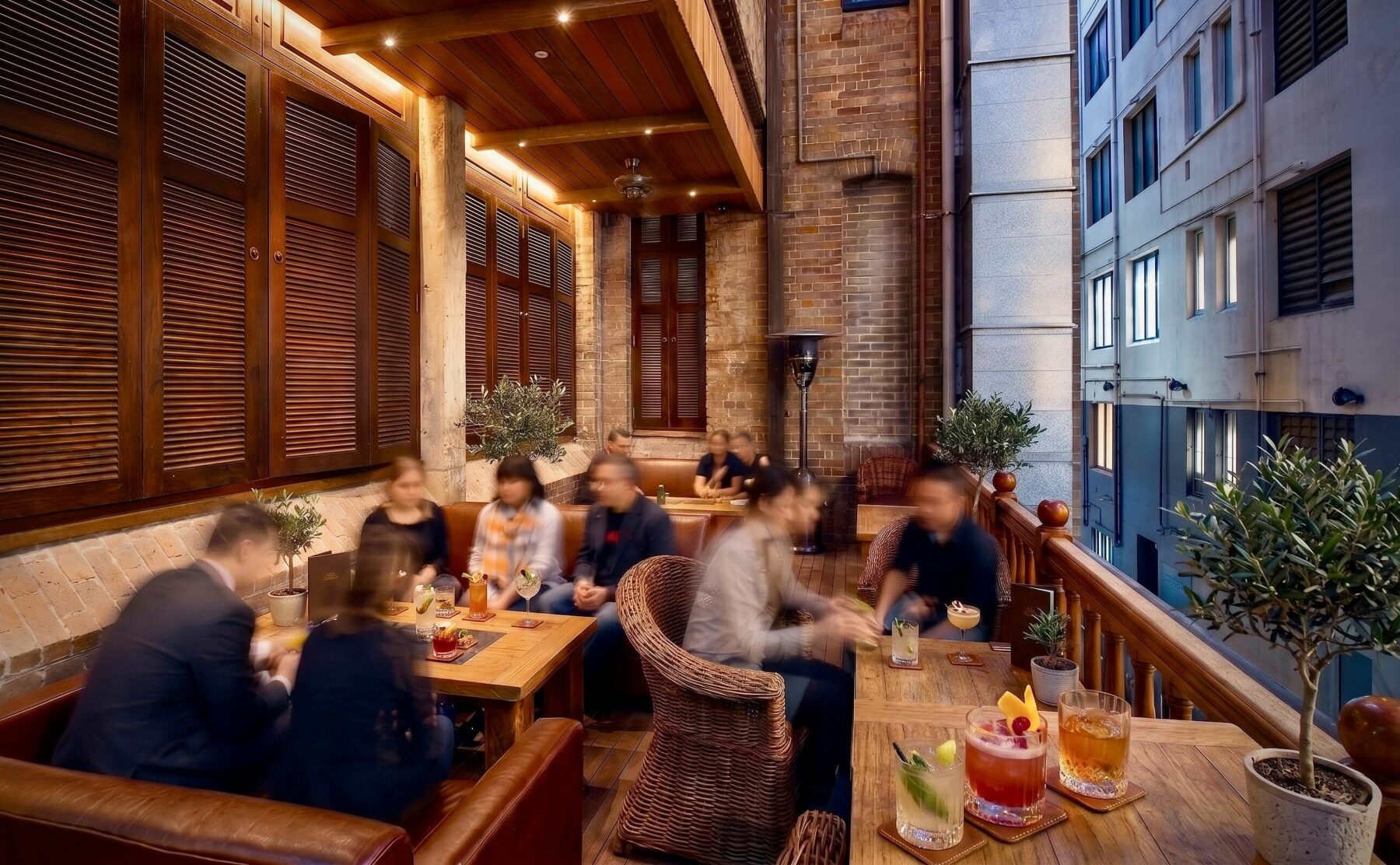 Heritage balcony lounge, timber window shutters, exposed brickwork wall, timber balustrade railing, wicker club chairs, linear timber tables, outdoor hospitality lighting, laneway view.
