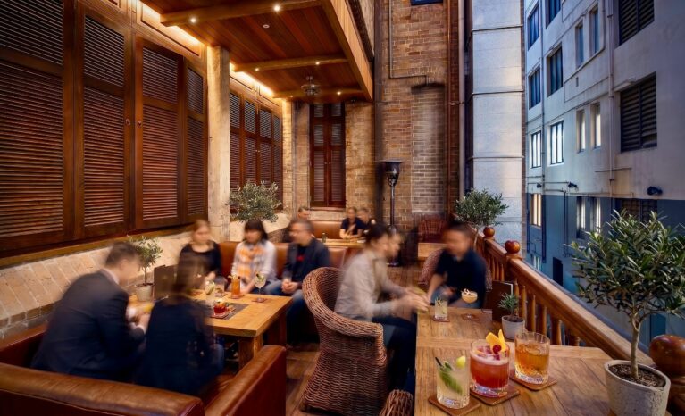 Heritage balcony lounge, timber window shutters, exposed brickwork wall, timber balustrade railing, wicker club chairs, linear timber tables, outdoor hospitality lighting, laneway view.