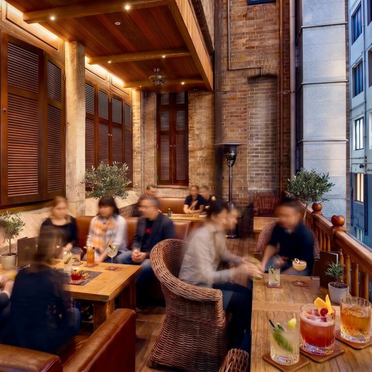 Heritage balcony lounge, timber window shutters, exposed brickwork wall, timber balustrade railing, wicker club chairs, linear timber tables, outdoor hospitality lighting, laneway view.