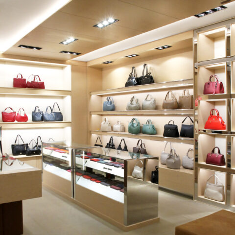 Bespoke retail shelving, luxury handbag display, maple veneer joinery, integrated LED strip lights, glass counter showcase, polished steel trims, recessed ceiling downlights, premium retail layout.