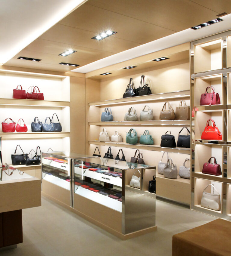Bespoke retail shelving, luxury handbag display, maple veneer joinery, integrated LED strip lights, glass counter showcase, polished steel trims, recessed ceiling downlights, premium retail layout.