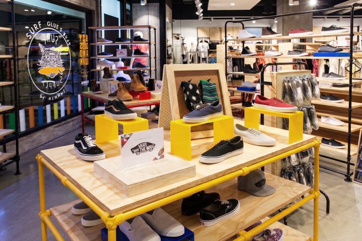 A retail shoe display unit showcasing vibrant yellow scaffolding frames, plywood shelving plinths, open-plan footwear racking, and polished concrete commercial flooring.