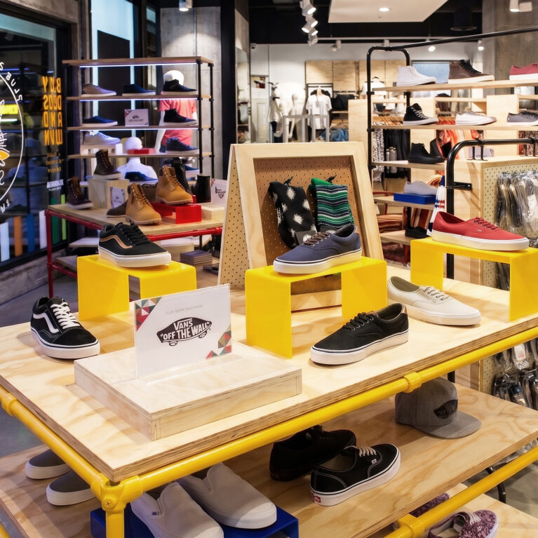 A retail shoe display unit showcasing vibrant yellow scaffolding frames, plywood shelving plinths, open-plan footwear racking, and polished concrete commercial flooring.