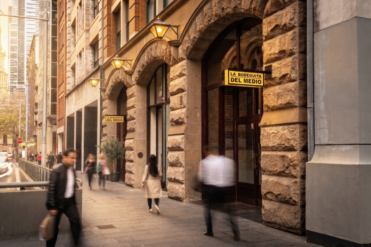 Heritage sandstone arches, historic building facade, custom cantilevered signage, vintage wall lanterns, timber entry doors, commercial street entrance, architectural streetscape, stone masonry portal.