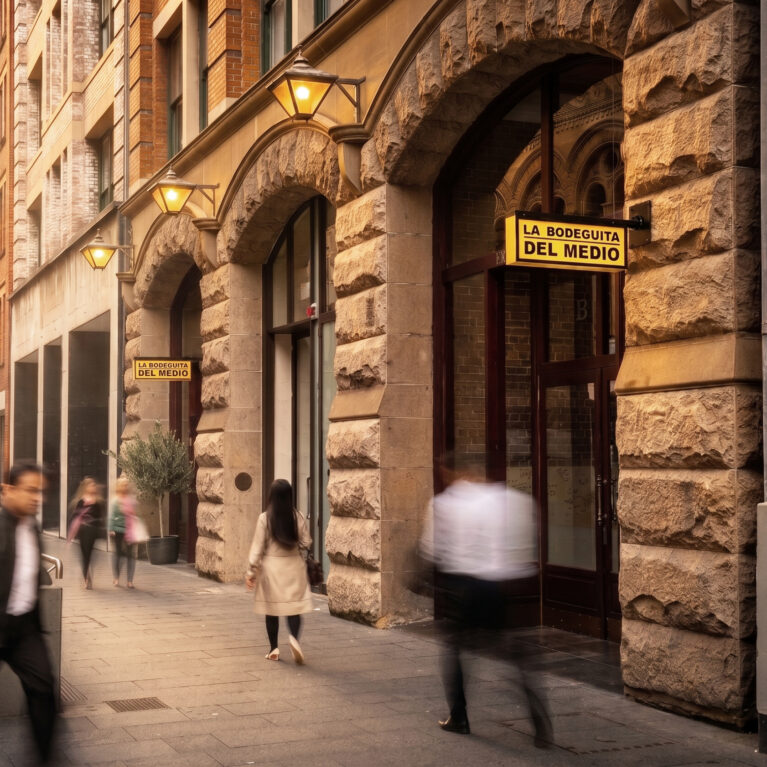 Heritage sandstone arches, historic building facade, custom cantilevered signage, vintage wall lanterns, timber entry doors, commercial street entrance, architectural streetscape, stone masonry portal.