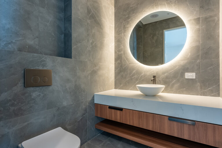 A modern vanity in the powder room exudes sophistication with a backlit circular mirror, sleek stone walls, and warm wood accents, creating an inviting and elegant space.