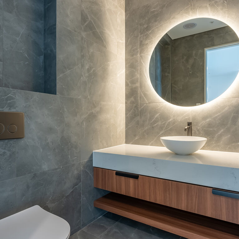 A modern vanity in the powder room exudes sophistication with a backlit circular mirror, sleek stone walls, and warm wood accents, creating an inviting and elegant space.