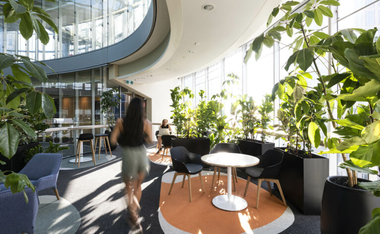 A light-filled workplace lounge framed by curved glazing and dense greenery, offering staff a quiet retreat for informal meetings, focused work or simply pausing with city views.
