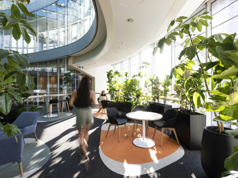 A light-filled workplace lounge framed by curved glazing and dense greenery, offering staff a quiet retreat for informal meetings, focused work or simply pausing with city views.
