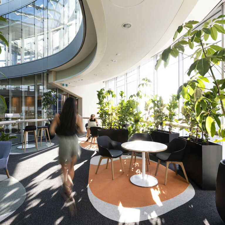 A light-filled workplace lounge framed by curved glazing and dense greenery, offering staff a quiet retreat for informal meetings, focused work or simply pausing with city views.