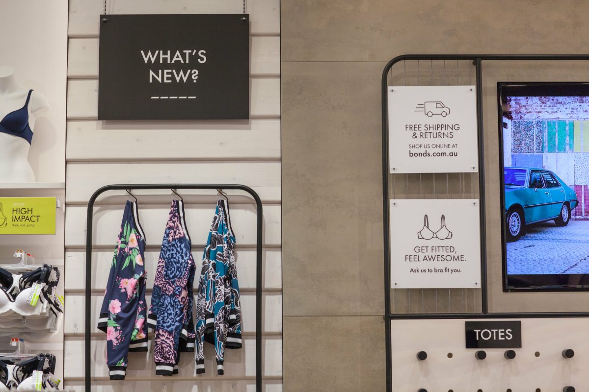 Retail merchandise display, horizontal wall paneling, matte black hanging rails, digital signage screens, promotional retail graphics, pegboard storage, modern store fit-out.