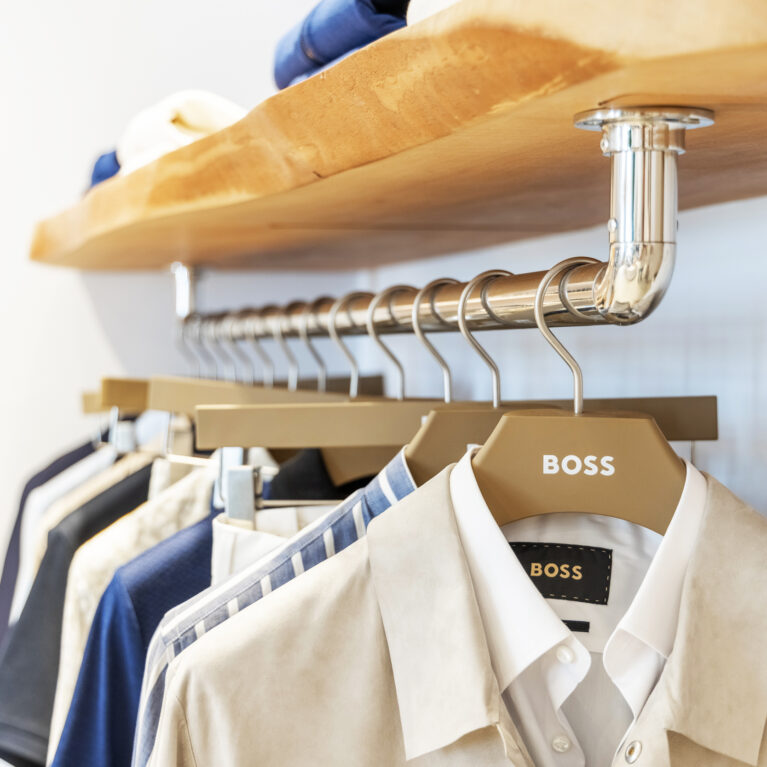 The clothing display balances sophistication and natural elements, featuring solid wood shelves, polished metal fixtures, and custom-branded hangers. The blend of organic textures and sleek finishes enhances the boutique’s contemporary yet relaxed aesthetic.