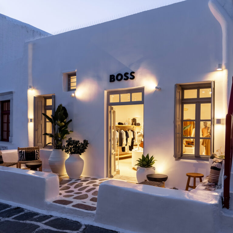 The store’s whitewashed façade comes alive at dusk, with warm lighting spilling onto the cobbled pathway. Thoughtful illumination highlights architectural features, creating an inviting contrast between the softly lit interior and the deepening blue of the evening sky.