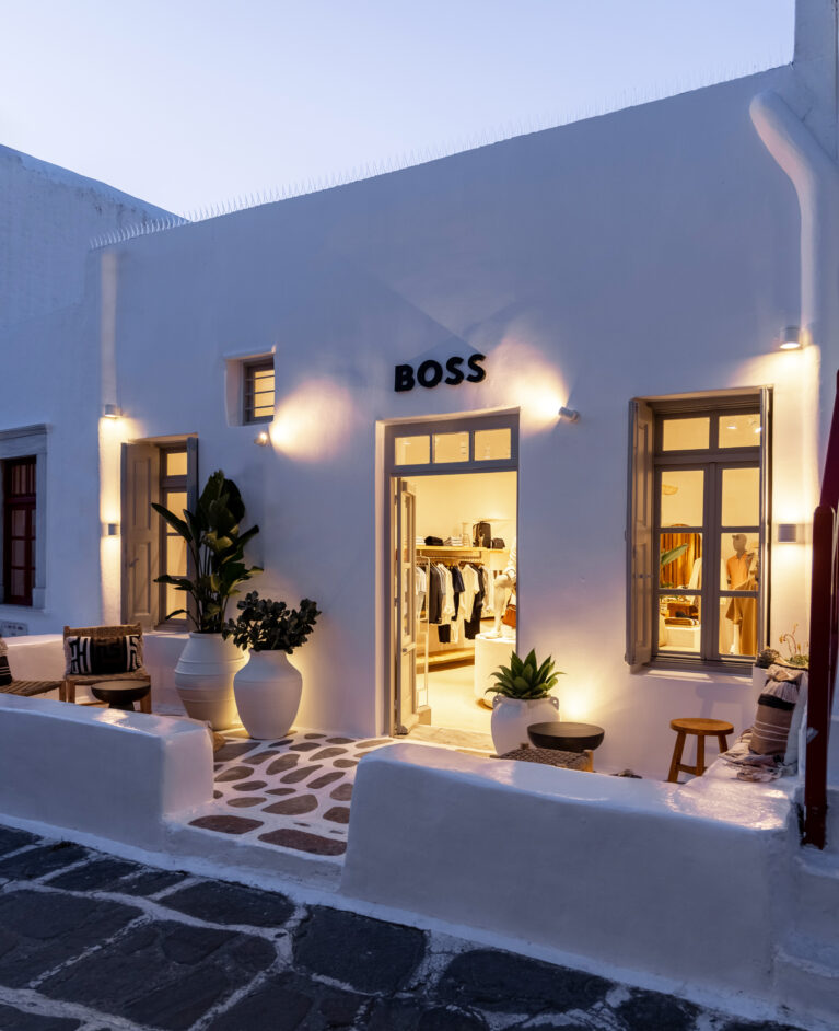 The store’s whitewashed façade comes alive at dusk, with warm lighting spilling onto the cobbled pathway. Thoughtful illumination highlights architectural features, creating an inviting contrast between the softly lit interior and the deepening blue of the evening sky.