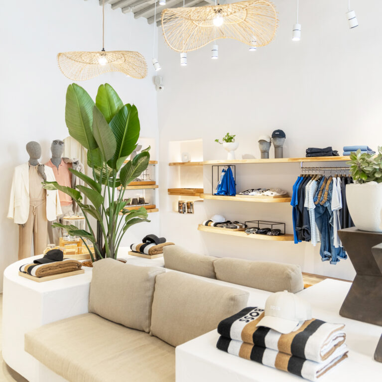 This inviting retail space blends modern elegance with island-inspired comfort. Soft linen seating, organic wooden shelving, and lush greenery create a calming atmosphere, while woven pendant lights enhance the airy aesthetic with a touch of artisanal craftsmanship.