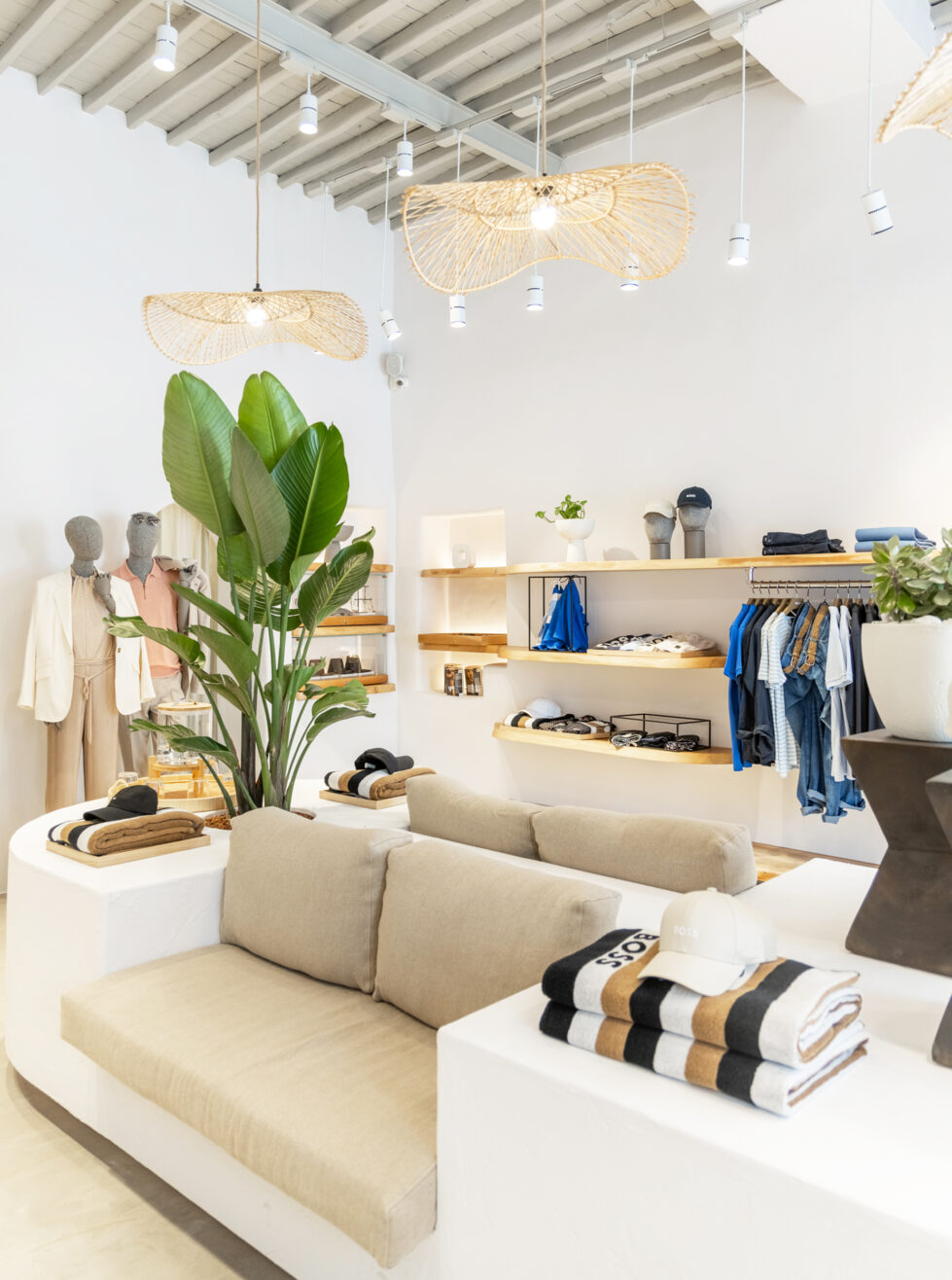 This inviting retail space blends modern elegance with island-inspired comfort. Soft linen seating, organic wooden shelving, and lush greenery create a calming atmosphere, while woven pendant lights enhance the airy aesthetic with a touch of artisanal craftsmanship.