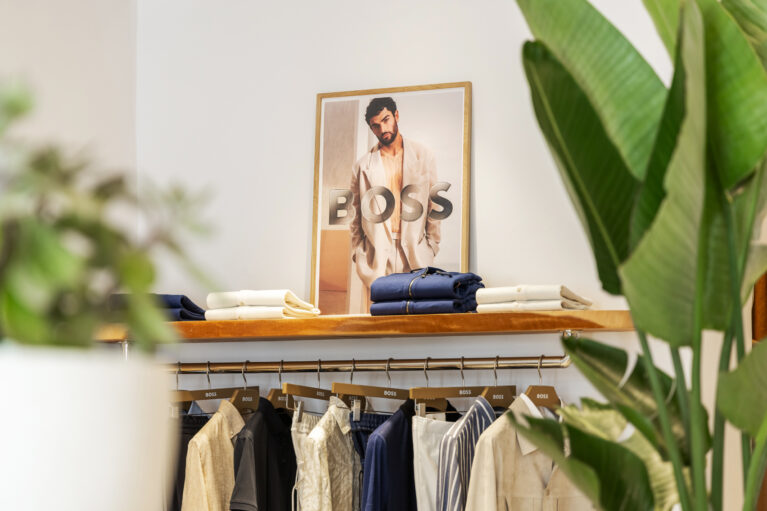 A curated selection of garments is displayed with precision, blending soft neutral tones and deep blues. The warm wood shelving and green foliage add an organic contrast, reinforcing the store’s effortlessly sophisticated and natural aesthetic.