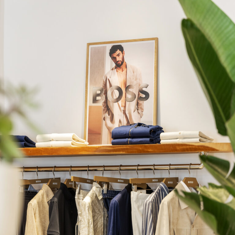 A curated selection of garments is displayed with precision, blending soft neutral tones and deep blues. The warm wood shelving and green foliage add an organic contrast, reinforcing the store’s effortlessly sophisticated and natural aesthetic.